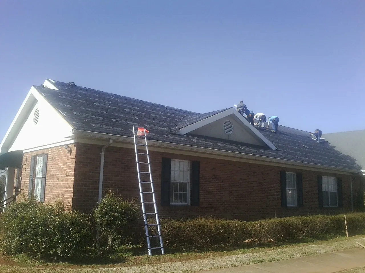 Crew tearing shingles off a brick ranch roof with a ladder set — Metal Roof Repair work by our crew in Dibble