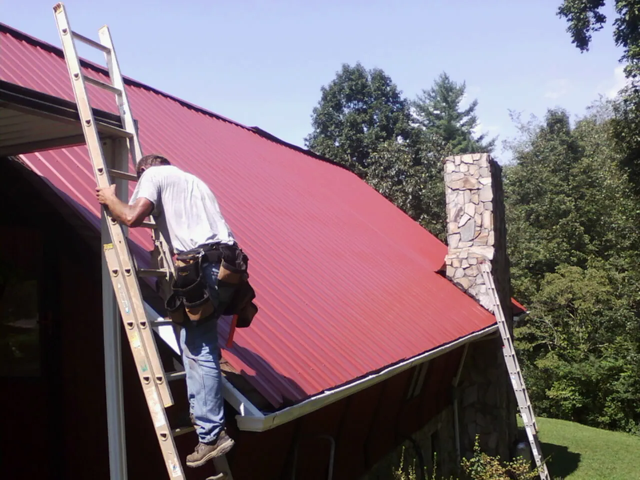 Roofer on a ladder beside a red metal roof and stone chimney — Soffit & Fascia Repair by Roofing Brothers in Dibble